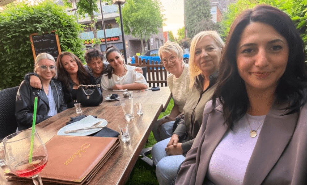 Seven women sit around a wooden outdoor cafe table, smiling at the camera during a sunny group photo.