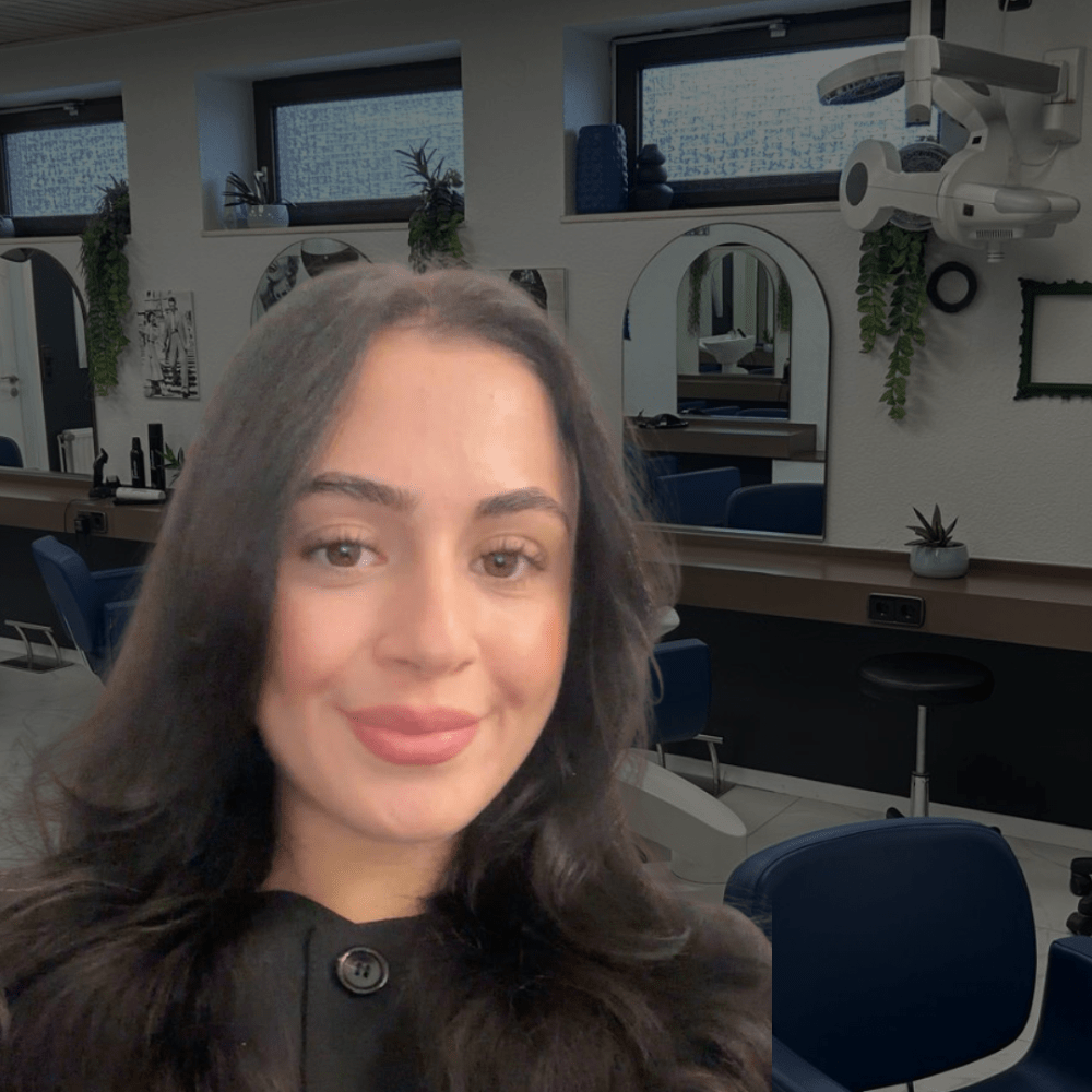 Close-up selfie of a smiling woman in a modern salon with blue chairs and mirrors in the background.